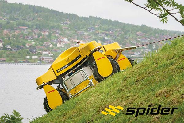 Ein gelber Spider Großflächen- und Böschungsmäher mäht mit Hilfe seiner Seilwinde einen steilen Hang. Im Hintergrund sind ein Fluss und am anderen Flussufer ein Dorf zu sehen. Ein gelber Spider Großflächen- und Böschungsmäher mäht mit Hilfe seiner Seilwinde einen steilen Hang. Im Hintergrund sind ein Fluss und am anderen Flussufer ein Dorf zu sehen.