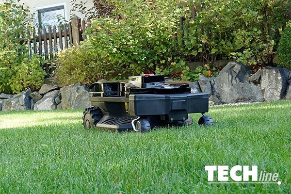 Ein schwarzer kleiner Roboterrasenmäher fährt auf einen gleichmäßig geschnittenen Rasen in einem Garten. Der Garten ist von einer Steinmauer, einem Zaun und Sträuchern begrenzt. Ein schwarzer kleiner Roboterrasenmäher fährt auf einen gleichmäßig geschnittenen Rasen in einem Garten. Der Garten ist von einer Steinmauer, einem Zaun und Sträuchern begrenzt.